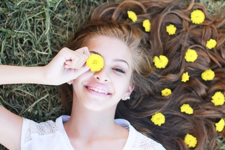 Girl laying on a ground with dandelions in her hair and smiles while putting a dandelion to her eye.